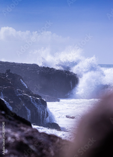 Fotografia vertical da água do mar a embater com força na rocha, espuma branca das ondas com sol e céu azul