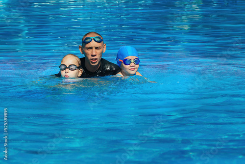 Swimming coach conducts a lesson with two girls in an outdoor pool with blue water on a warm sunny day