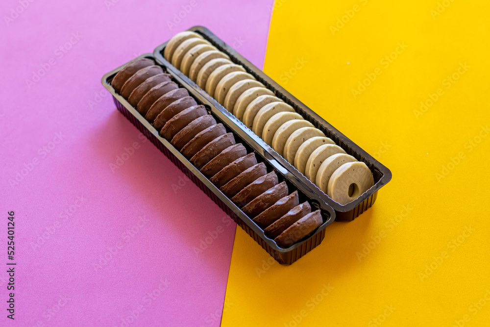 Angled view of small donuts covered in white and dark chocolate, in plastic cases, on a combined pink and yellow background. Angled view of small donuts covered in white and dark chocolate, in plastic cases, on a combined pink and yellow background.