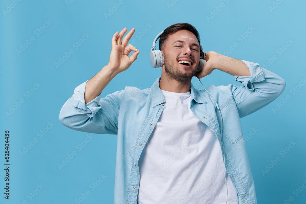 Happy attractive tanned handsome man in casual basic t-shirt headphones listen cool sound show gesture Okay posing isolated on blue studio background. Copy space Banner Mockup. Music concept Happy attractive tanned handsome man in casual basic t-shirt headphones listen cool sound show gesture Okay posing isolated on blue studio background. Copy space Banner Mockup. Music concept