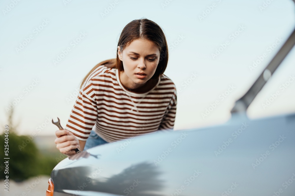 A woman with a wrench looks sadly and sadly under the open hood of her car and can't fix it from a roadside breakdown while traveling alone