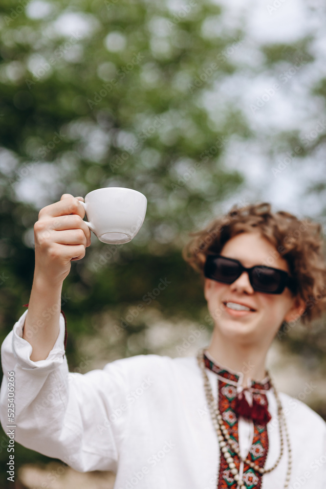 Naklejka premium A curly blond guy in an embroidered shirt and black glasses holds a cup of coffee in his hands.Coffee love concept.