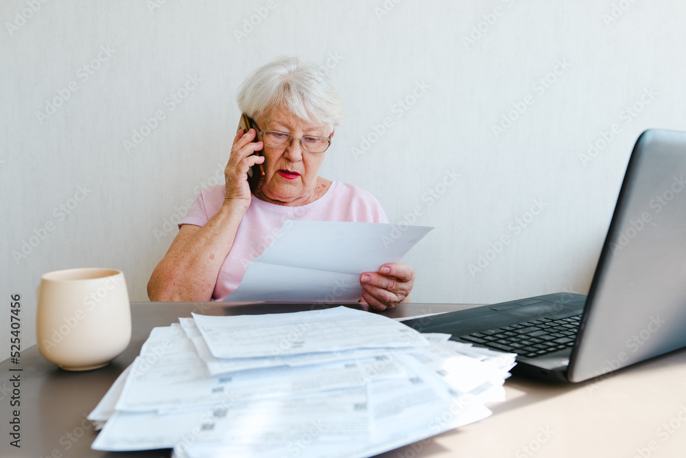 Senior woman holding papers busy at laptop managing house utility bills or finances, aged female using computer working with bank loan or mortgage documents online. Elderly Senior woman holding papers busy at laptop managing house utility bills or finances, aged female using computer working with bank loan or mortgage documents online. Elderly