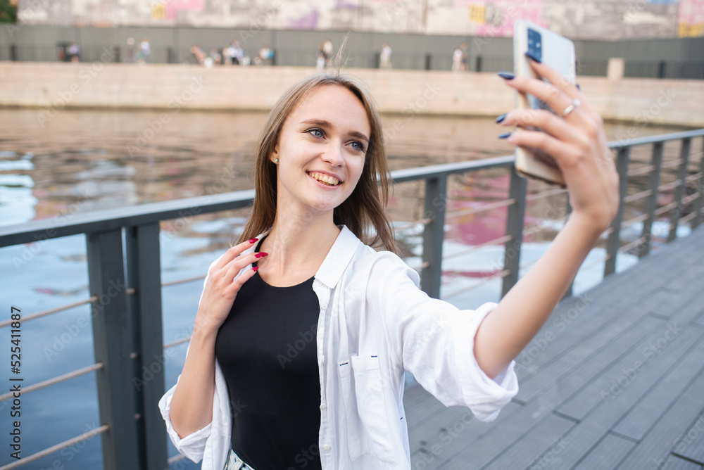A young and attractive Caucasian girl in casual clothes takes a selfie on a mobile phone camera in a park near the river.