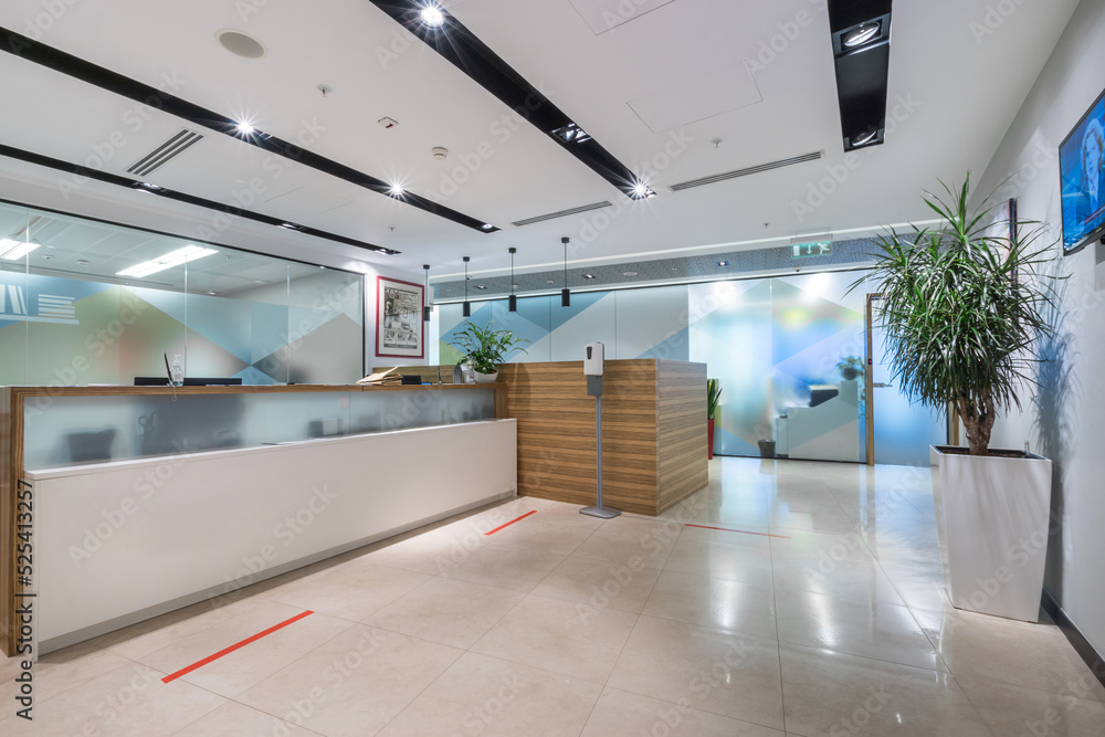 A brightly lit spacious office lobby with glass walls and a reception desk. Red line markings for social distance