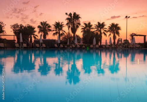 Beautiful reflection in swimming pool at colorful sunset. Purple sky reflected in water, palm trees, sun beds, umbrellas at night in summer. Luxury resort. Landscape with empty pool in twilight