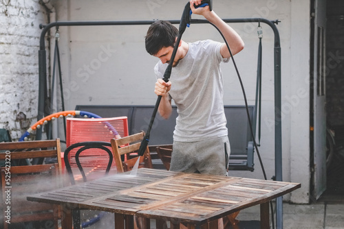 A caucasian guy in gray sweatpants and a T-shirt washes a wooden garden table with a karcher photo