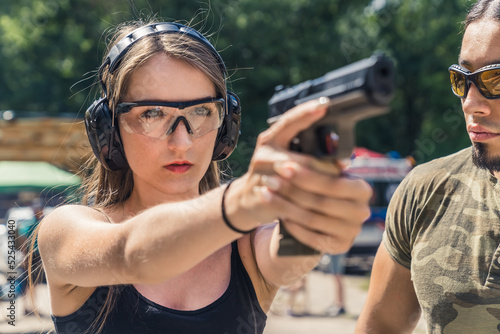 Caucasian young woman wearing protective goggles and headphones aiming handgun observed by male bearded instructor. Firearms training. Horiozntal shot. High quality photo