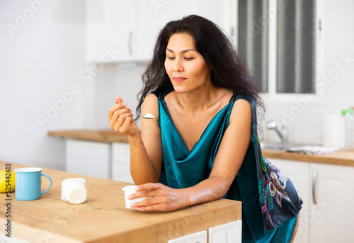 Portrait of asian woman in nightie eating yoghurt in kitchen at home. Woman leaning on kitchen table  eating yogurt.