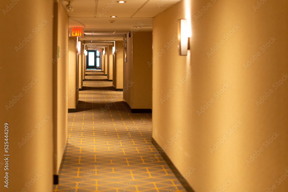A long narrow hallway of a hotel with a red exit sign light sconce lights and cream colored walls. The carpet on the floor is green and beige with a pattern. The low ceiling has white tiles. 