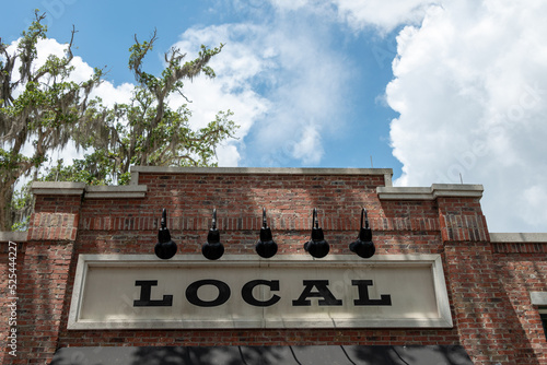 A cream colored informational sign with the word local in black lettering is affixed to a vintage red and brown colored brick building. There are multiple black barn lights hanging over the signage.  photo