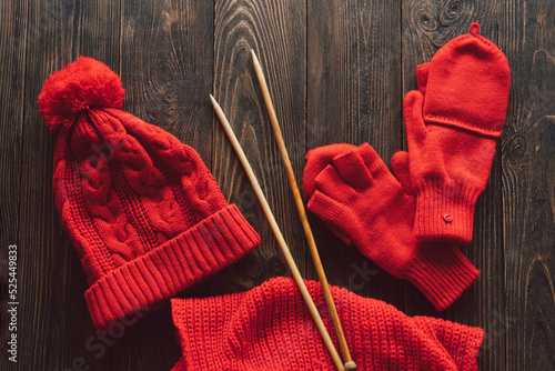 Knitted red hat, scarf, knitted mittens and wooden knitting needles on wooden background. Hobby craft. Concept of knitting, needlework.