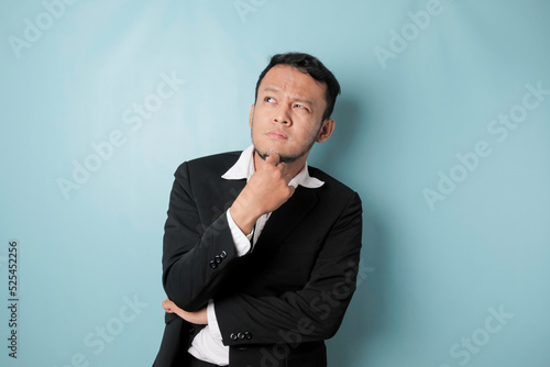 Portrait of a thoughtful young Asian businessman wearing black suit looking aside while his finger on his chin isolated over blue background