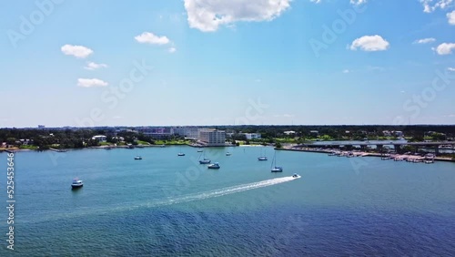 A causeway connecting Belleair Beach and Pinellas Beaches is surrounded by a clear, emerald-colored water, many boats, a clear sky, and a nearby township was captured from above by Drone photo