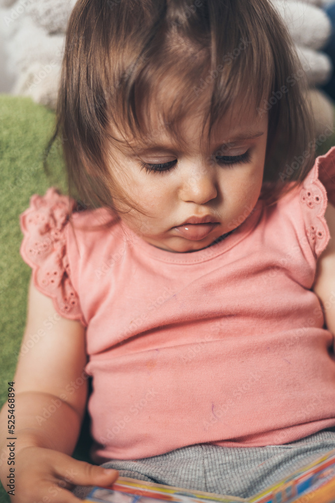 Pensive baby girl sitting on sofa at home, looking down with smart and curious beautiful hazel brown eyes. Childhood concept. Family concept. Pensive baby girl sitting on sofa at home, looking down with smart and curious beautiful hazel brown eyes. Childhood concept. Family concept.