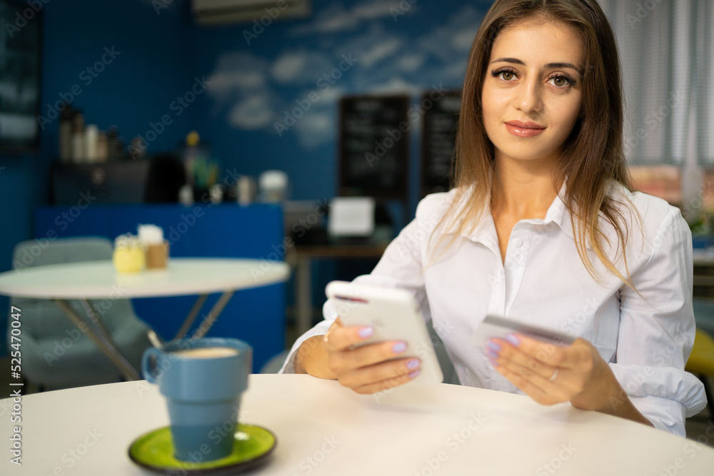 Beautiful young girl with a cheerful expression holding a smartphone and a credit card, banking online or shopping while in a cafe interior. payment and vacation Beautiful young girl with a cheerful expression holding a smartphone and a credit card, banking online or shopping while in a cafe interior. payment and vacation