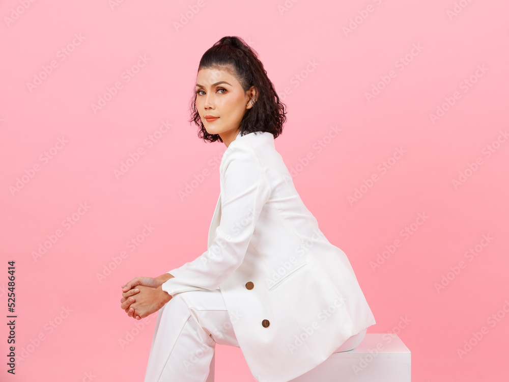 Portrait studio shot of Asian young sexy curly hairstyle professional successful businesswoman in white fashion casual suit sitting posing holding hands together on square box on pink background