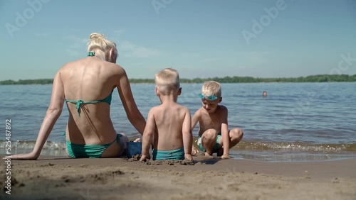 A young woman with her two sons is having a great vacation on the beach. Children are happy to be together and play with their mother. Mom shows them what figures can be made from sand. photo