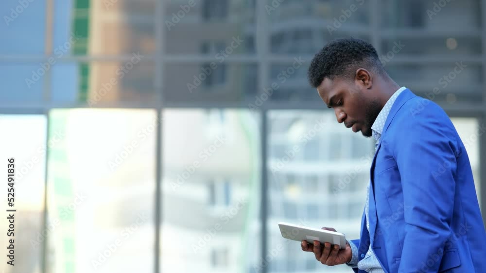 African American man holding white tablet walks past glass office building. Mature black businessman explores work of interesting device in street African American man holding white tablet walks past glass office building. Mature black businessman explores work of interesting device in street