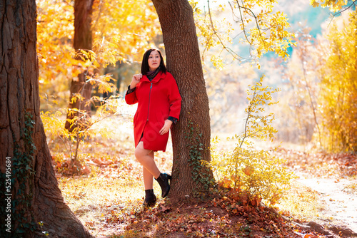 An elegantly dressed adult woman in a red coat poses coquettishly with her foot on a tree trunk. Autumn Park in the sunlight in the background photo