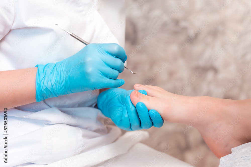Pedicurist does a pedicure for the client's foot, cleaning the nails with a double-sided curette. Close up. The concept of chiropody and podology Pedicurist does a pedicure for the client's foot, cleaning the nails with a double-sided curette. Close up. The concept of chiropody and podology