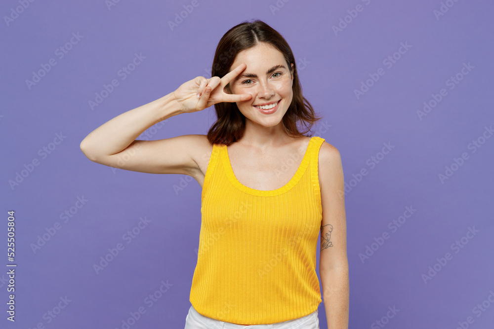 Young smiling caucasian fun cool woman 20s she wearing yellow tank shirt showing victory sign looking camera isolated on plain pastel light purple background studio portrait. People lifestyle concept. Young smiling caucasian fun cool woman 20s she wearing yellow tank shirt showing victory sign looking camera isolated on plain pastel light purple background studio portrait. People lifestyle concept.