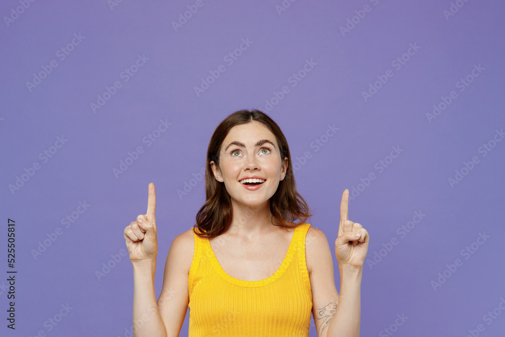 Young happy fun woman 20s wear yellow tank shirt point index finger overhead indicate on workspace area copy space mock up isolated on plain pastel light purple background. People lifestyle concept. Young happy fun woman 20s wear yellow tank shirt point index finger overhead indicate on workspace area copy space mock up isolated on plain pastel light purple background. People lifestyle concept.