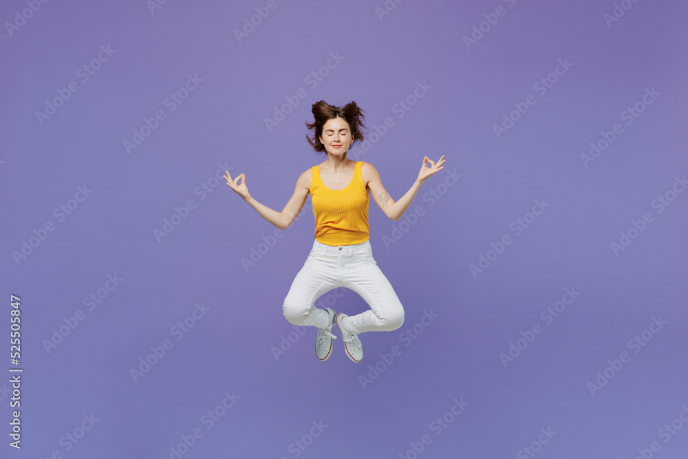 Full body young spiritual tranquil woman wear yellow tank shirt jump high hold spreading hands in yoga om aum gesture relax meditate try to calm down isolated on plain pastel light purple background.
