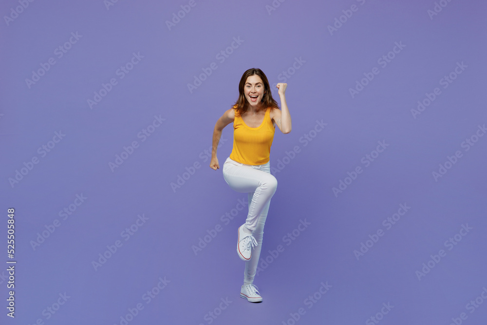 Full body young smiling happy woman 20s she wear yellow tank shirt look camera clench fist run fast hurry up isolated on plain pastel light purple background studio portrait. People lifestyle concept.