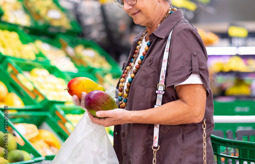 Senior woman holding two fresh mango in a supermarket or grocery store close-up. Woman holds two mature mangos wearing protective gloves photo