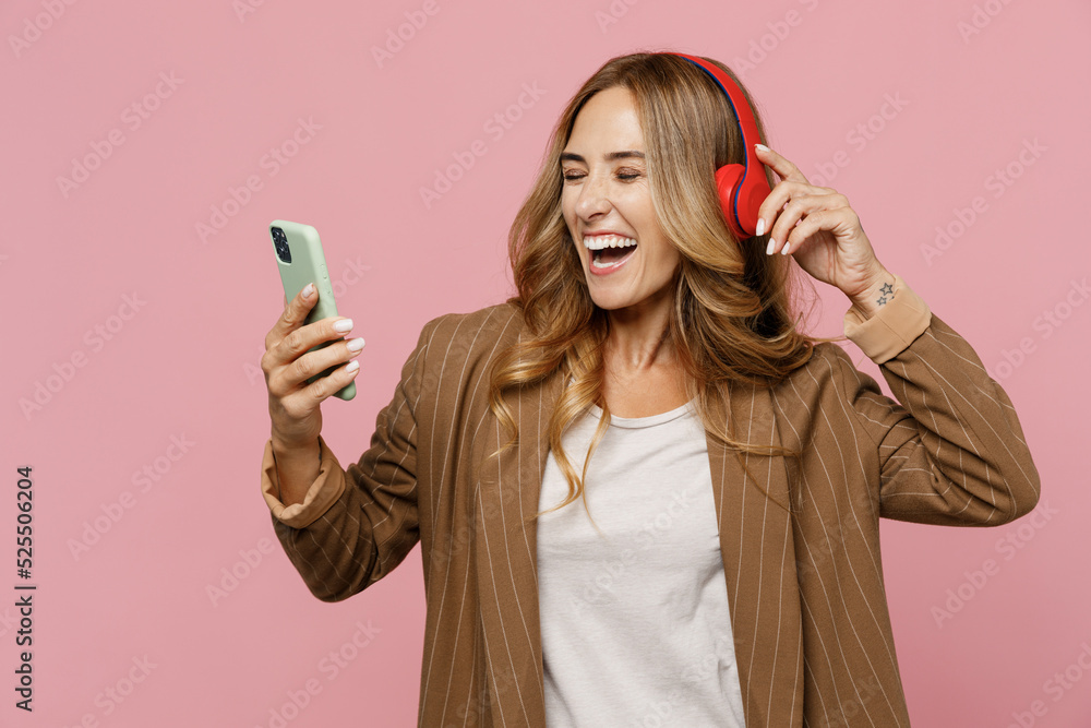 Young fun successful employee business woman 30s she wear casual classic jacket headphones sing song in microphone at karaoke club use mobile cell phone isolated on plain pastel light pink background. Young fun successful employee business woman 30s she wear casual classic jacket headphones sing song in microphone at karaoke club use mobile cell phone isolated on plain pastel light pink background.