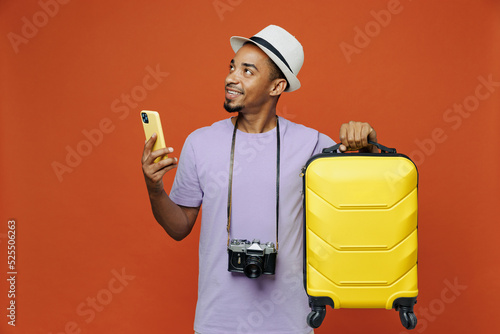 Traveler black man wear purple t-shirt hat hold suitcase mobile phone isolated on plain orange color background. Tourist travel abroad on weekends spare time getaway. Air flight trip journey concept. #525506263