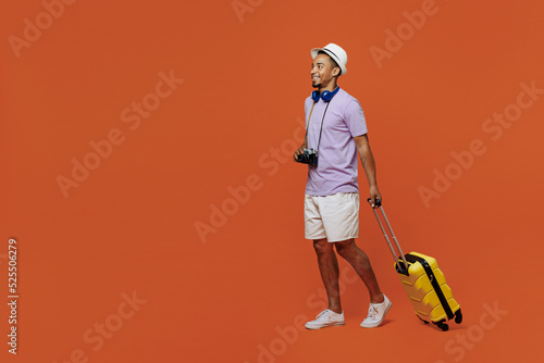 Full body traveler black man wear purple t-shirt hat headphones go with suitcase isolated on plain orange background. Tourist travel abroad in spare time rest getaway. Air flight trip journey concept. #525506279