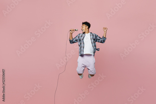 Full body young smiling man of African American ethnicity he wear blue shirt sing song in microphone jump high isolated on plain pastel light pink background studio portrait. People lifestyle concept.