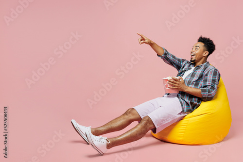 Fotografiet Full body young man of African American ethnicity wear blue shirt sit in bag chair hold popcorn bucket point finger aside watch film isolated on plain pastel pink background