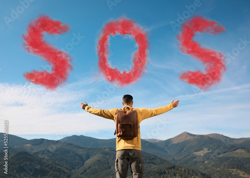 Man with backpack and word SOS made of color smoke bomb in mountains, back view