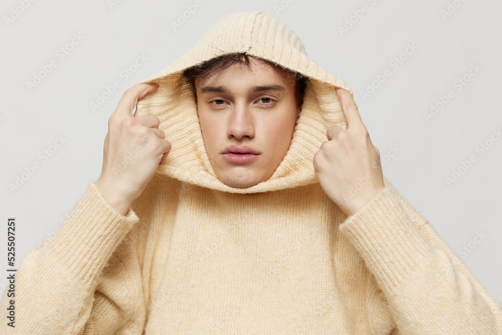 Horizontal studio shot. A handsome young man with dark short hair combed back in a beige sweater with a high neck stands on a gray background and pulls it over his head Horizontal studio shot. A handsome young man with dark short hair combed back in a beige sweater with a high neck stands on a gray background and pulls it over his head
