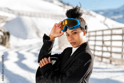 African American woman with goggles and a snowboard on a snowy mountain during winter
