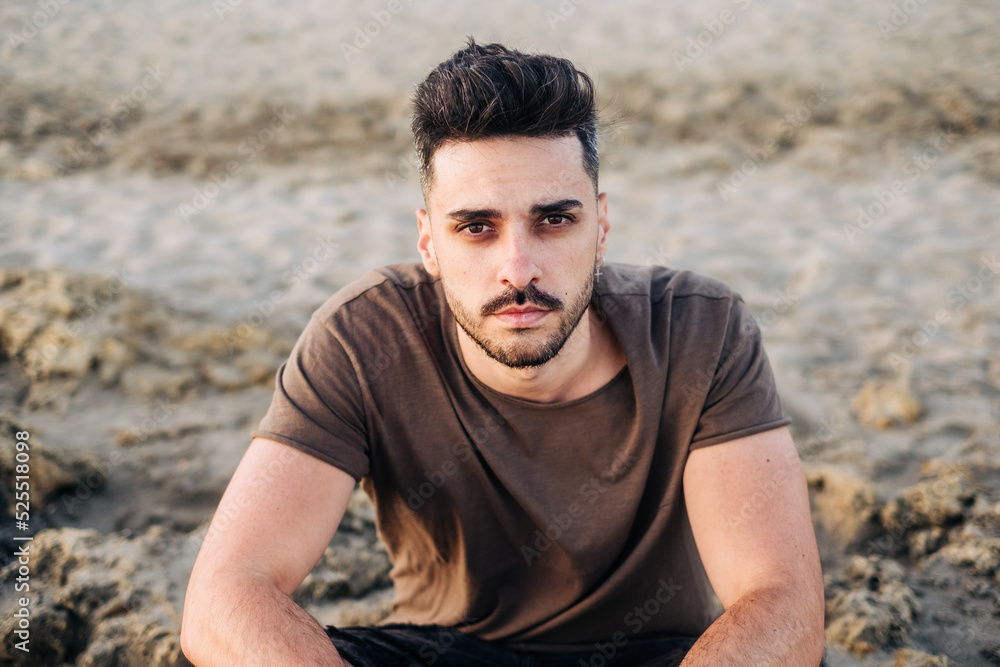 Handsome young Hispanic man sitting on sandy beach clasping hand Handsome young Hispanic man sitting on sandy beach clasping hand