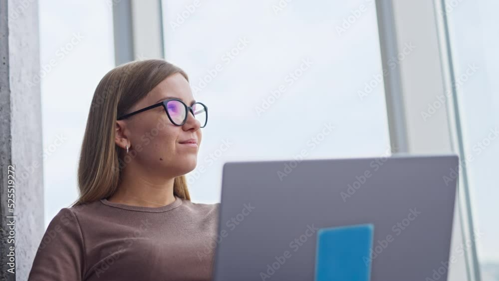 Confident relaxed blonde woman working on computer. Female looks into window for a moment and continues her work smiling. Low angle view. Confident relaxed blonde woman working on computer. Female looks into window for a moment and continues her work smiling. Low angle view.