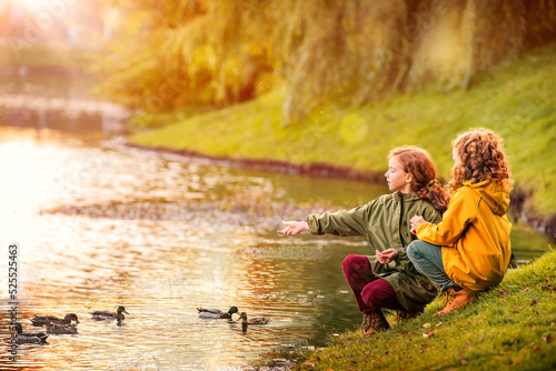 Two red-haired schoolgirl girls, sisters, cheerfully feeding ducks on the bank of a pond in a city park during the golden autumn leaf fall.