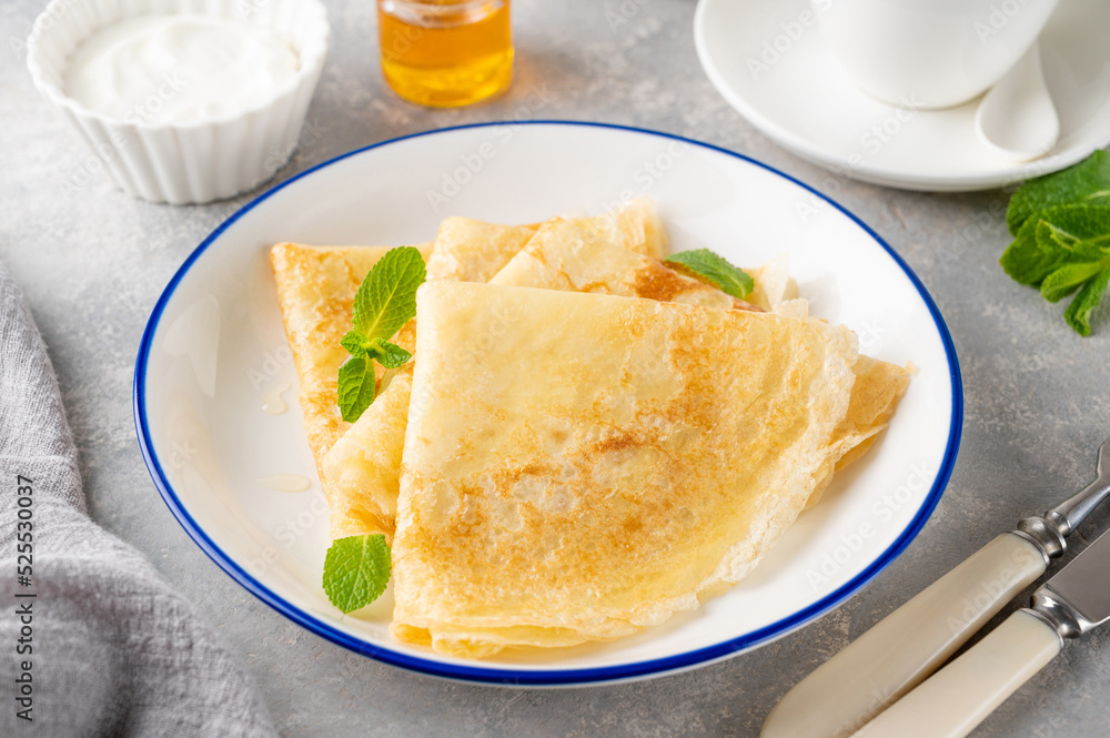 Stack of traditional thin pancakes or crepes on a plate with honey and sour cream on a gray concrete background. Food for Maslenitsa. Copy space