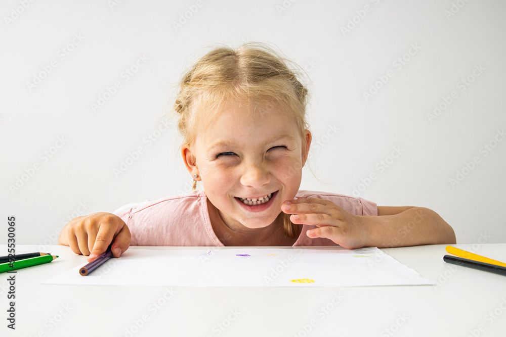 A smiling child blonde girl draws with colored pencils sitting at a white table A smiling child blonde girl draws with colored pencils sitting at a white table