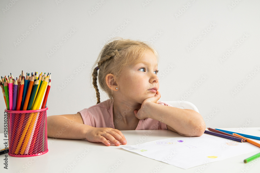 A thoughtful child blonde girl draws with colored pencils sitting at a white table A thoughtful child blonde girl draws with colored pencils sitting at a white table