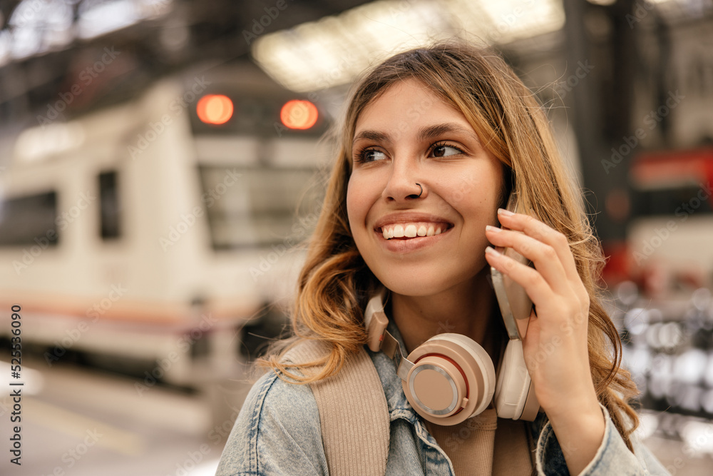 Cute young caucasian girl calls on mobile phone against background of locomotive. Blonde woman with wavy hair smiles teeth. Audio technology, gadgets concept Cute young caucasian girl calls on mobile phone against background of locomotive. Blonde woman with wavy hair smiles teeth. Audio technology, gadgets concept