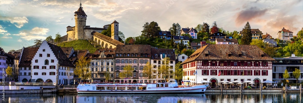 Fototapeta premium Historic Munot castle and medieval town of Schaffhausen in Switzerland reflected in Rhine river at sunset. 16K panoarama of old town architecture over calm water in evening light