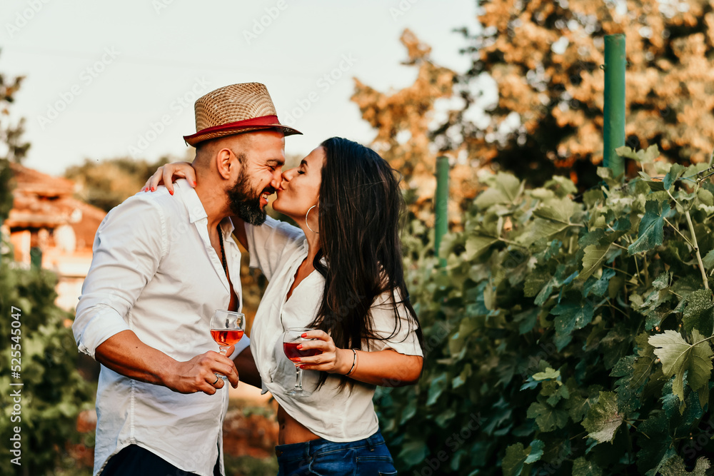 Portrait of a smiling  happy couple kissing    in a Vineyard toasting wine. Beautiful  brunette woman and bearded muscular man spending time together during grape harvest.