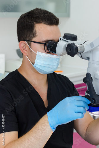 A young dentist with black hair looks through a microscope. He is wearing gloves and a medical mask