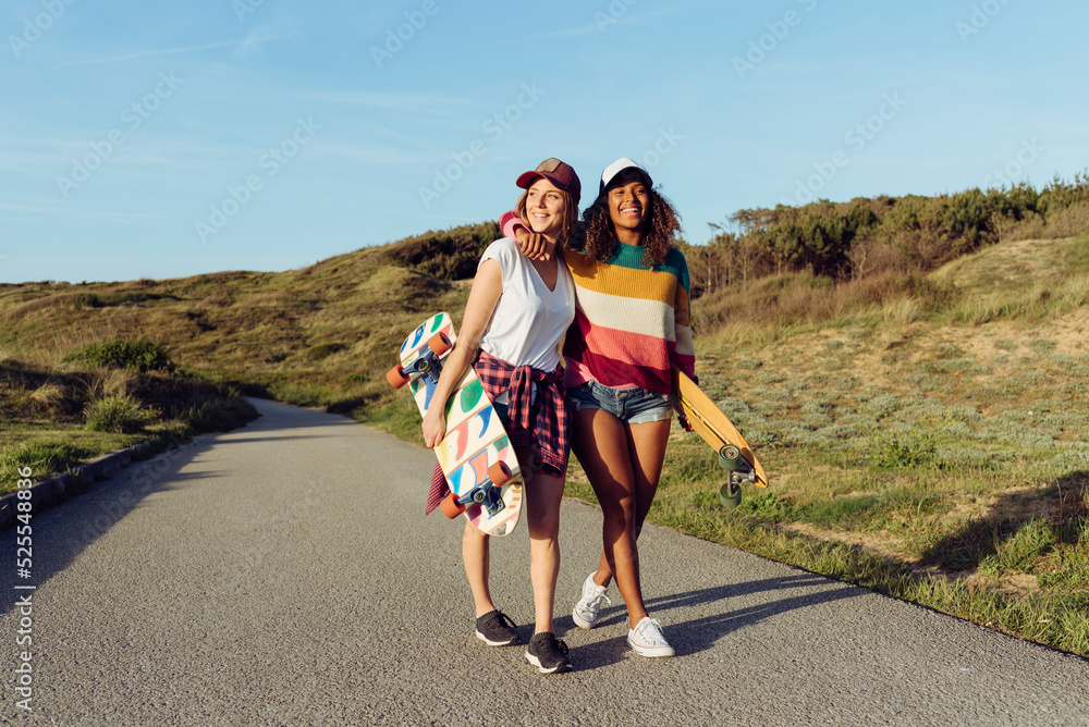 Women friends walking down the road holding skate boards Women friends walking down the road holding skate boards