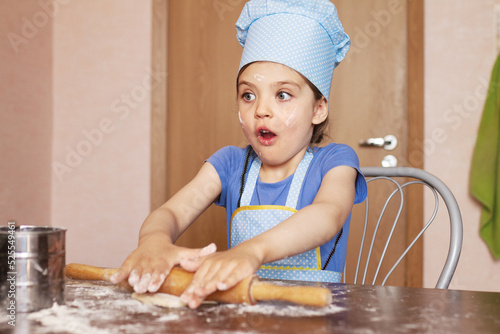 5 year old surprised girl in blue dress and dressed as chef in kitchen laughs and rolling out dough